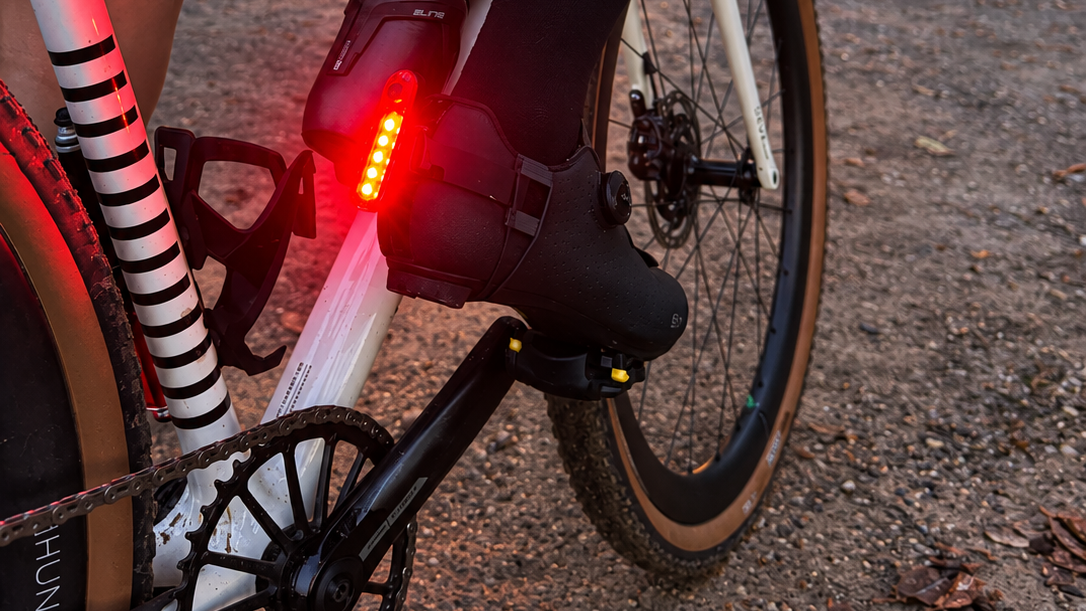 A close-up outdoor shot of a cyclist riding a white road bike at sunset, wearing a black cycling boot with an integrated red LED tail light mounted on cyclist shoe.