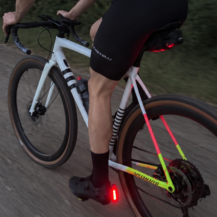 A cyclist wearing black cycling shorts and a black shirt rides a white gravel bike on a dirt road at dusk. The bike frame has neon yellow and pink accents, and a red light is visible on the cyclist heel.
